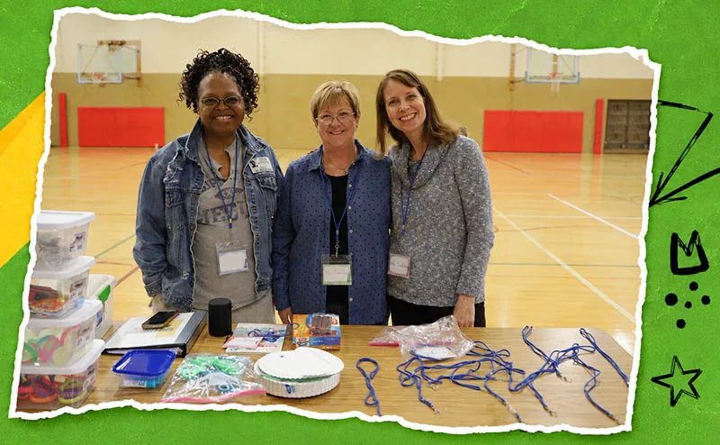 program-details Three club volunteers stand behind a table of activity supplies in a school gym.